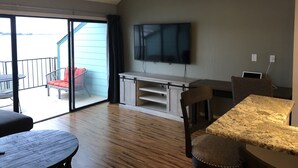 Un espacio habitable amplio con un televisor grande, piso de madera y un balcón con una silla y una mesa.