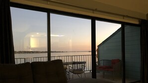 Una sala de estar con sofá y mesa, un balcón con una silla, y vista a un cuerpo de agua y un edificio lejano.