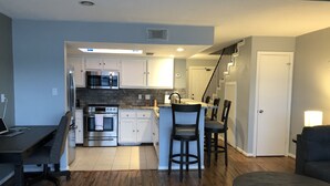 Una cocina moderna con electrodomésticos de acero inoxidable, un comedor con mesa y sillas de madera oscura, y una escalera que sube a otro nivel.