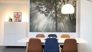 Uma zona de refeições moderna com uma grande obra de arte pendurada na parede retratando uma cena de floresta, uma mesa branca e cadeiras castanhas.