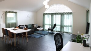 Uma sala de estar moderna com área de jantar, janelas amplas com cortinas, um sofá preto, uma mesa de centro e uma cadeira preta.