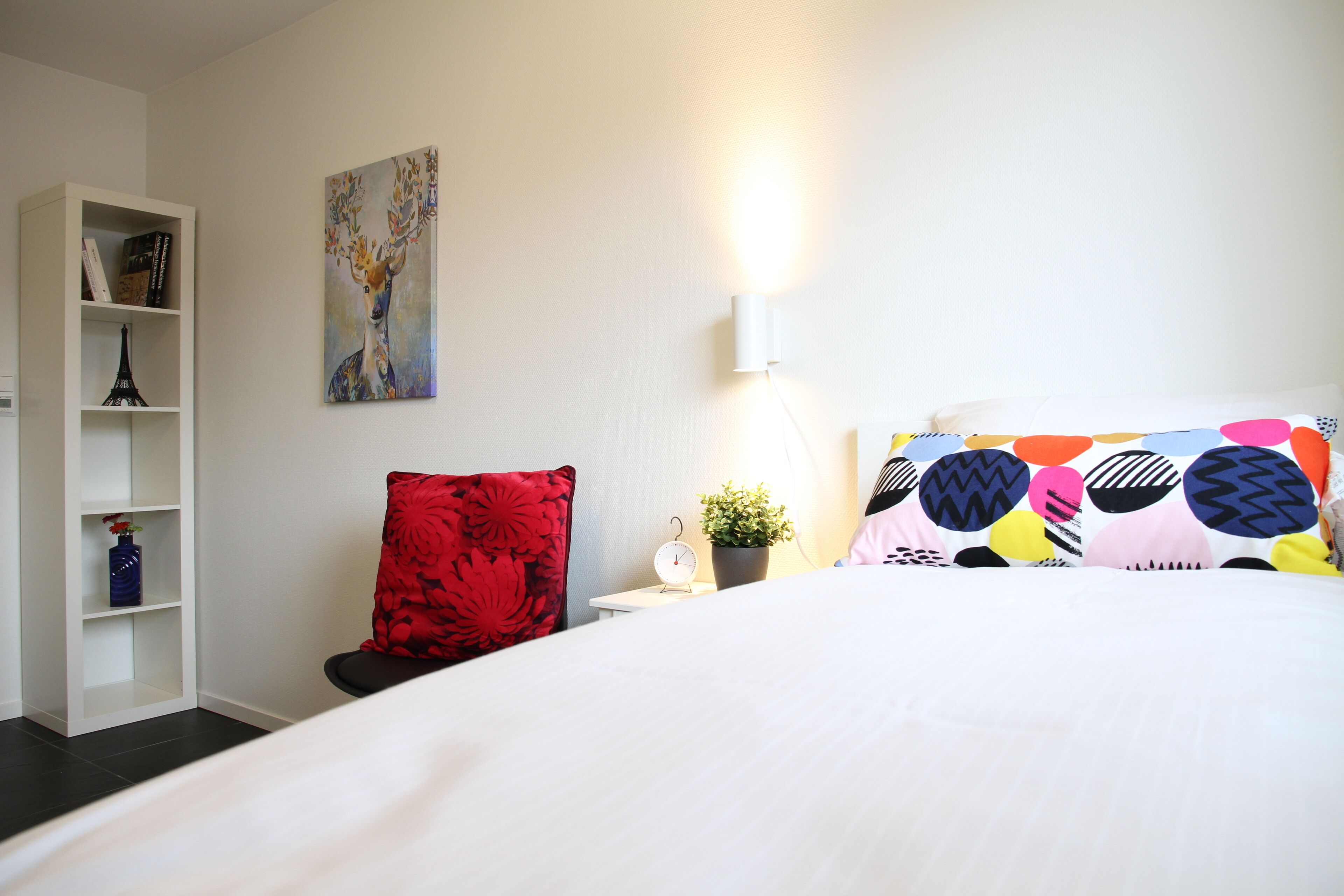 Habitación de hotel con una cama, una estantería, una silla roja, un reloj de pared y una planta en maceta.