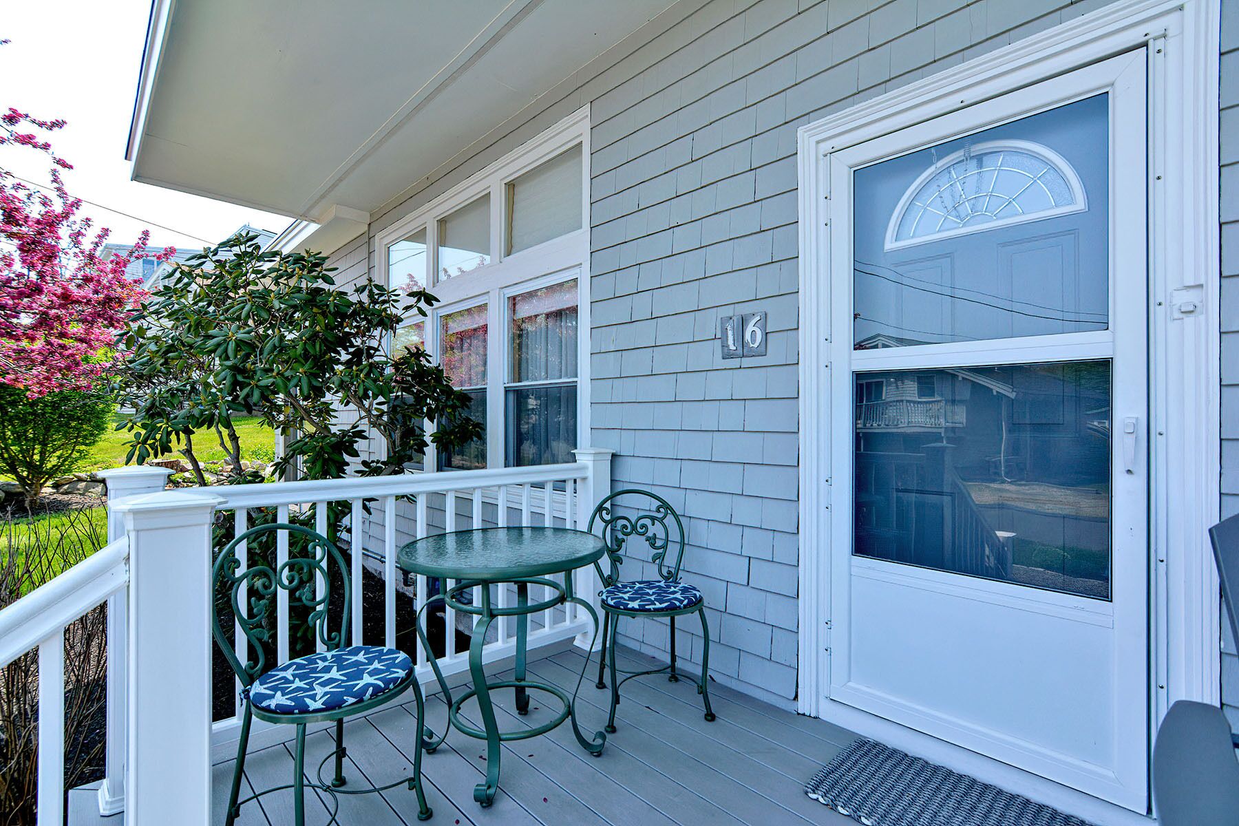 A porch with two green metal chairs and a small table, a white door with a window, and a house number 16.