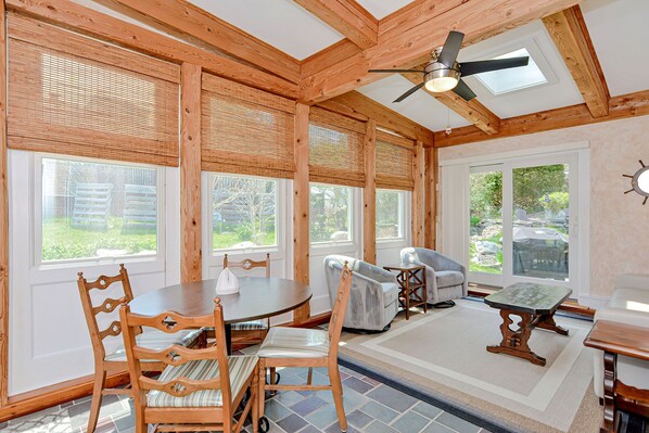 A room with wooden beams, a ceiling fan, a dining table with chairs, and large windows.