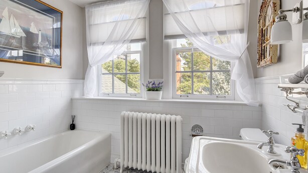 Ein Badezimmer mit einer Badewanne, einem Waschbecken, einem Fenster mit durchsichtigen Vorhängen, einem Heizkörper und einem gerahmten Bild an der Wand.