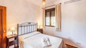 A neatly made bed with a white comforter and two towels, a bedside table with a lamp, a window with curtains, and a chandelier. - La Pedrosa Setenil de las Bodegas by Ruralidays (Cádiz)