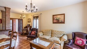 A living room with a fireplace, a chandelier, a sofa, a coffee table, and a dining area with chairs and a table. - La Pedrosa Setenil de las Bodegas by Ruralidays (Cádiz)