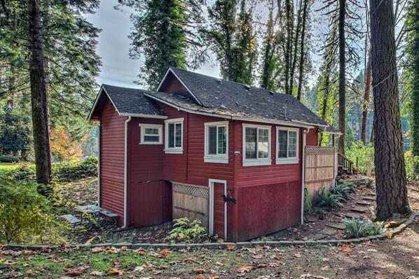 A small, single-story cabin with a red exterior and white trim, surrounded by tall trees and a forested area.