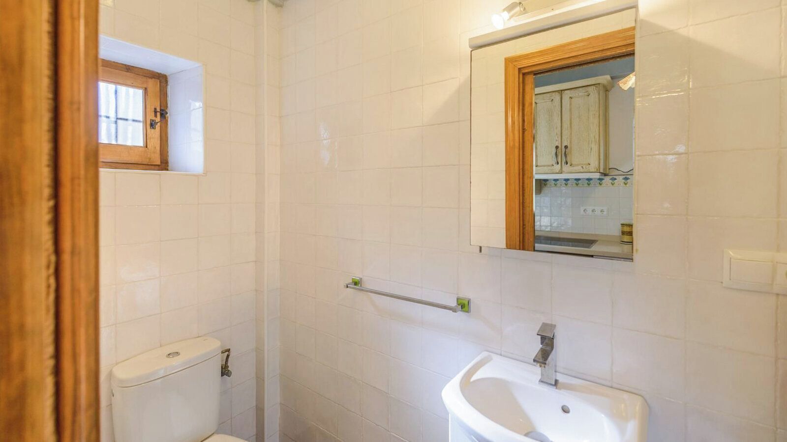 A bathroom with white tiles, a toilet, a sink, a mirror, and a window.
