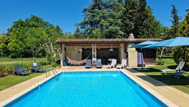 Piscina con sillones de descanso y sombrillas, rodeada de follaje y un edificio con techo de tejas.