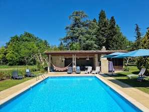 A swimming pool with lounge chairs and umbrellas, surrounded by greenery and a building with a tiled roof.