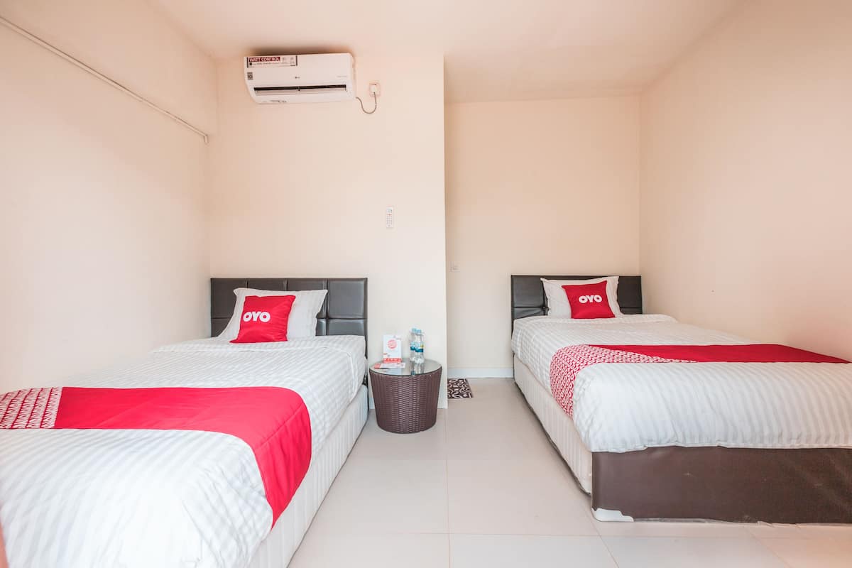 A hotel room with two beds, each with a red OYO pillow, a small wicker side table with a bottle and a card, and a wall-mounted air conditioner.