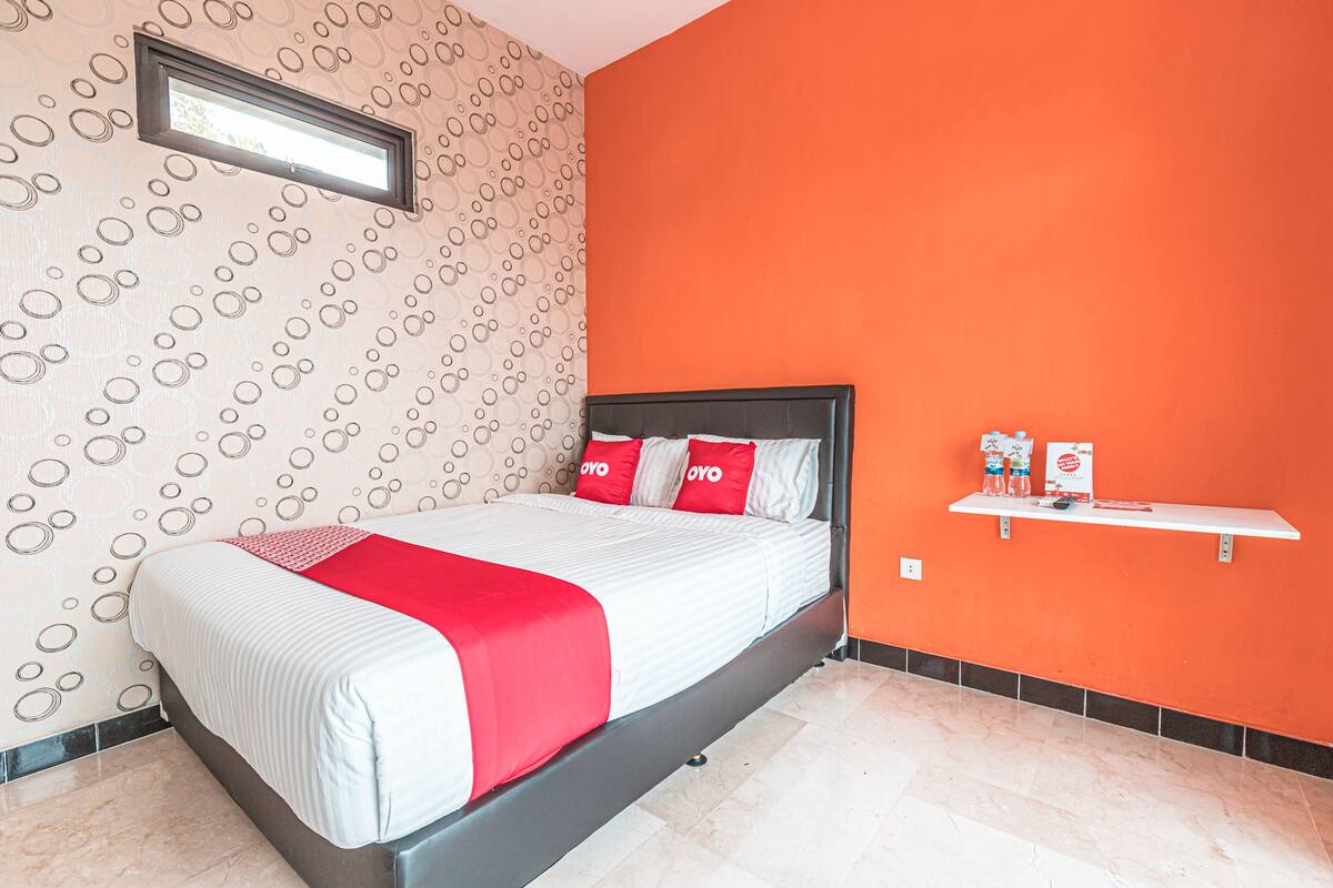 A hotel room with a bed, a red and white bedspread, a red pillow, a small shelf with items, and a patterned wallpaper.