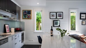 Une cuisine moderne avec des meubles blancs, un îlot central et un coin repas doté d’une table en verre. Les murs sont ornés d’œuvres d’art encadrées, et une fenêtre offre une vue sur la verdure.