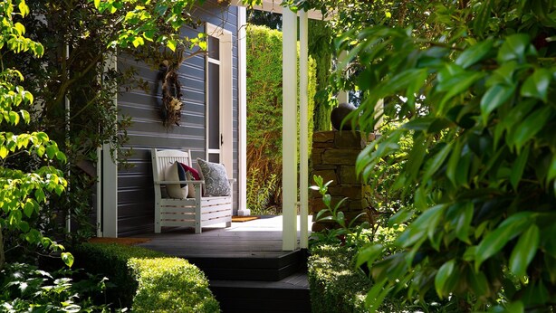 Une terrasse en bois avec un banc blanc et des coussins, entourée d’une végétation luxuriante et d’un mur de pierre.