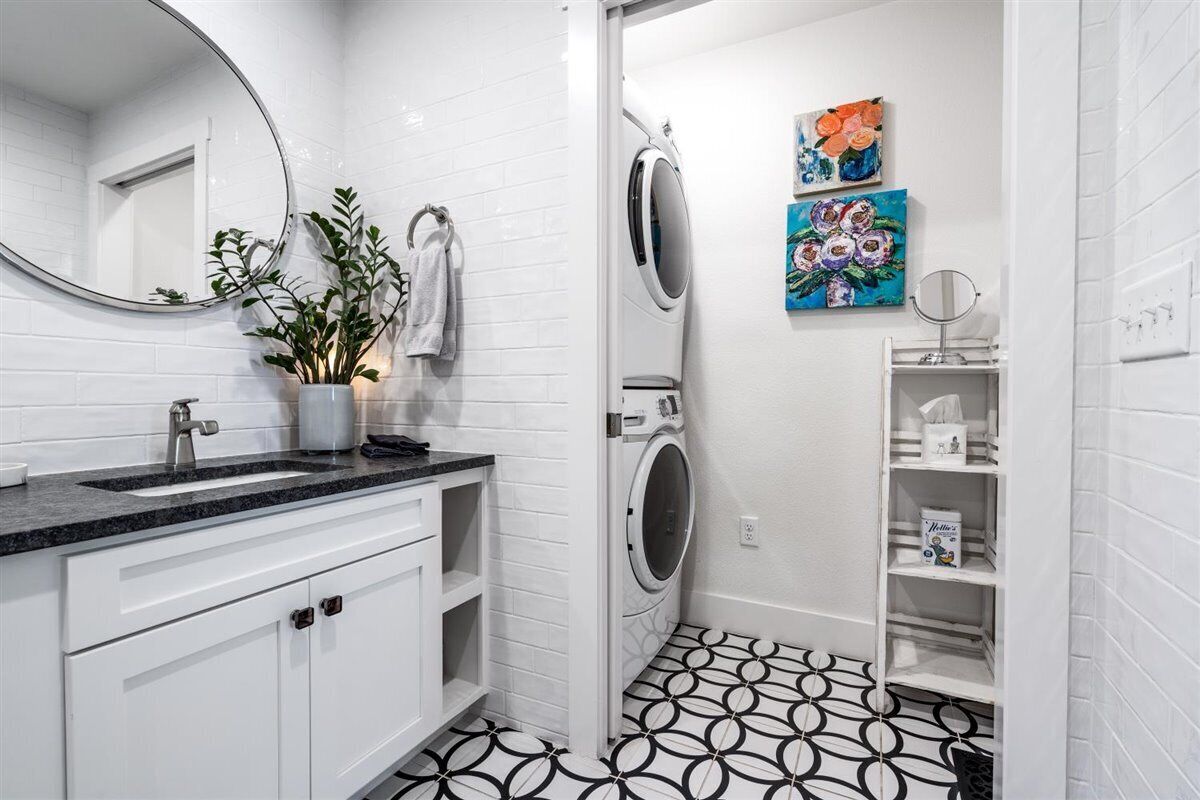 Un bagno moderno con parete rivestita in piastrelle bianche, pavimento a motivi bianchi e neri, lavandino con rubinetto, uno specchio, una lavatrice e due quadri colorati appesi al muro.