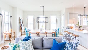 Un salon lumineux et moderne, avec un canapé d’angle gris, des coussins à motifs bleus et un coin repas comprenant une table et des chaises blanches.