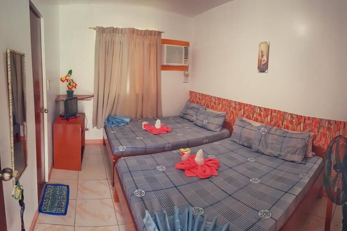 A room with two beds, a small desk with a computer, a window with curtains, and a wall-mounted air conditioner.