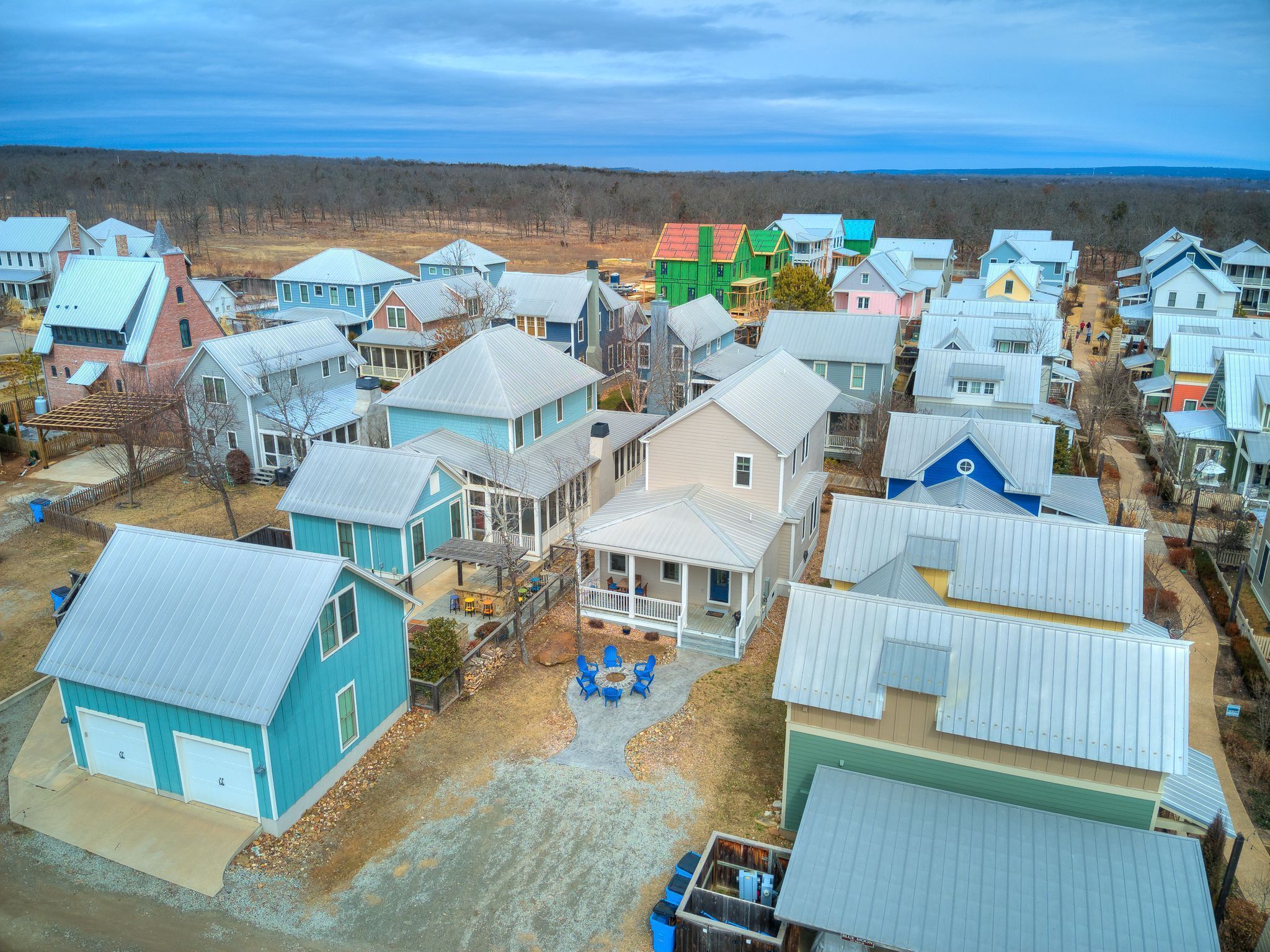 Un quartier résidentiel où les maisons présentent différents styles et couleurs de toits, une place centrale pavée avec des espaces de repos extérieurs, et un paysage boisé qui s’étend au loin.