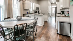 A modern kitchen with white cabinets, a central island, and a dining area with a round table and chairs. - Carlton Landing! Charming Cottage on boardwalk! (Carlton Landing)