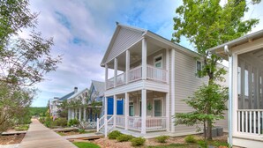 A row of white two-story houses with balconies, a wooden walkway, and a garden area with shrubs and a small tree. - Carlton Landing! Charming Cottage on boardwalk! (Carlton Landing)