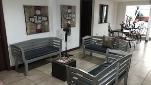 Una sala de estar con dos sofás grises, una mesa de centro con libros, un ventilador de pie y un comedor con sillas y una mesa.