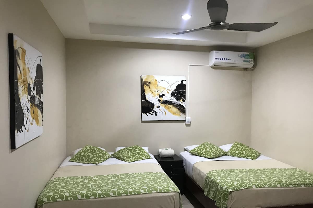 Habitación de hotel con dos camas individuales, cada una con ropa de cama de diseño verde, ventilador de techo, equipo de aire acondicionado y obras de arte abstractas en las paredes.