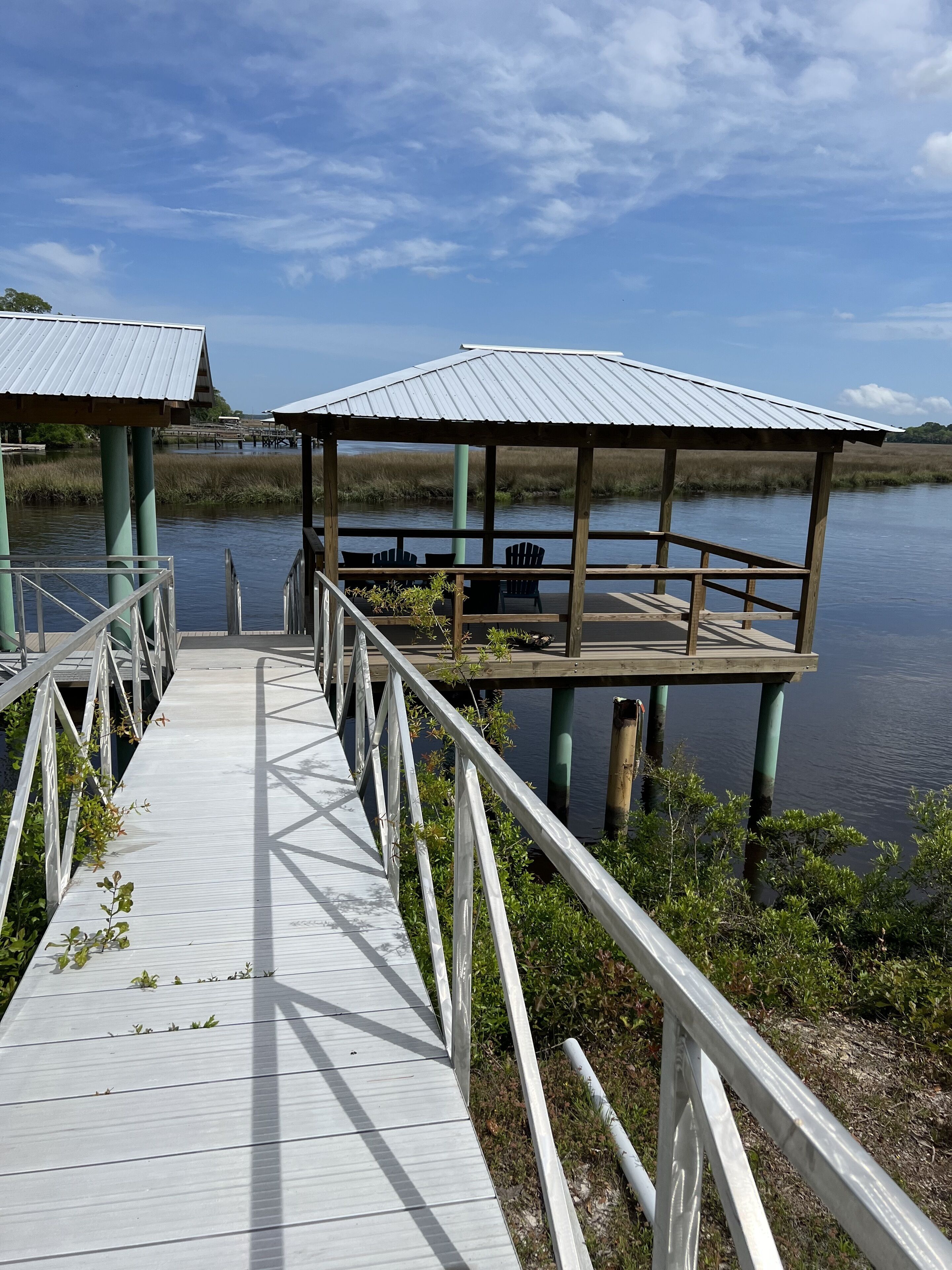 A wooden boardwalk leading to a covered pavilion with chairs, surrounded by water and vegetation.