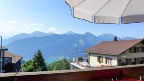 Un balcon avec une table rouge et un parasol blanc, d’où l’on peut admirer une chaîne de montagnes et un lac.
