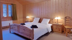 Une chambre à coucher aux murs lambrissés, avec un lit simple, deux tables de chevet avec des lampes, une chaise et une fenêtre avec des stores.