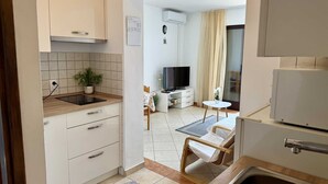 A compact kitchen with white cabinets, a built-in oven, and a sink. There's a small dining area with a table and chairs, a TV, and a plant. - Apartment Gordana 1 with sea view (Omisalj)