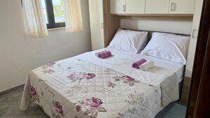A neatly made bed with floral bedding and rolled towels, a window with curtains, and a wardrobe with a light wood finish.