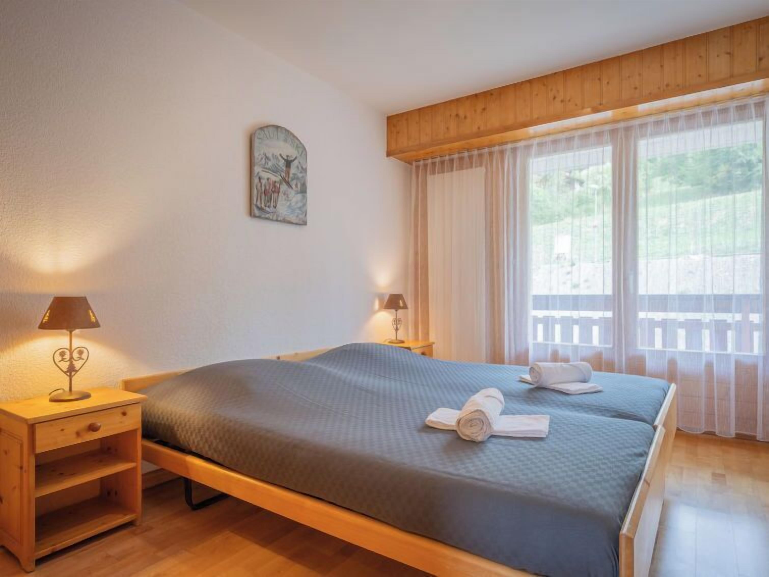Un lit double avec une couvre-lit bleue, deux lampes de chevet, une table de chevet en bois, un tableau au mur et une fenêtre avec des rideaux légers.