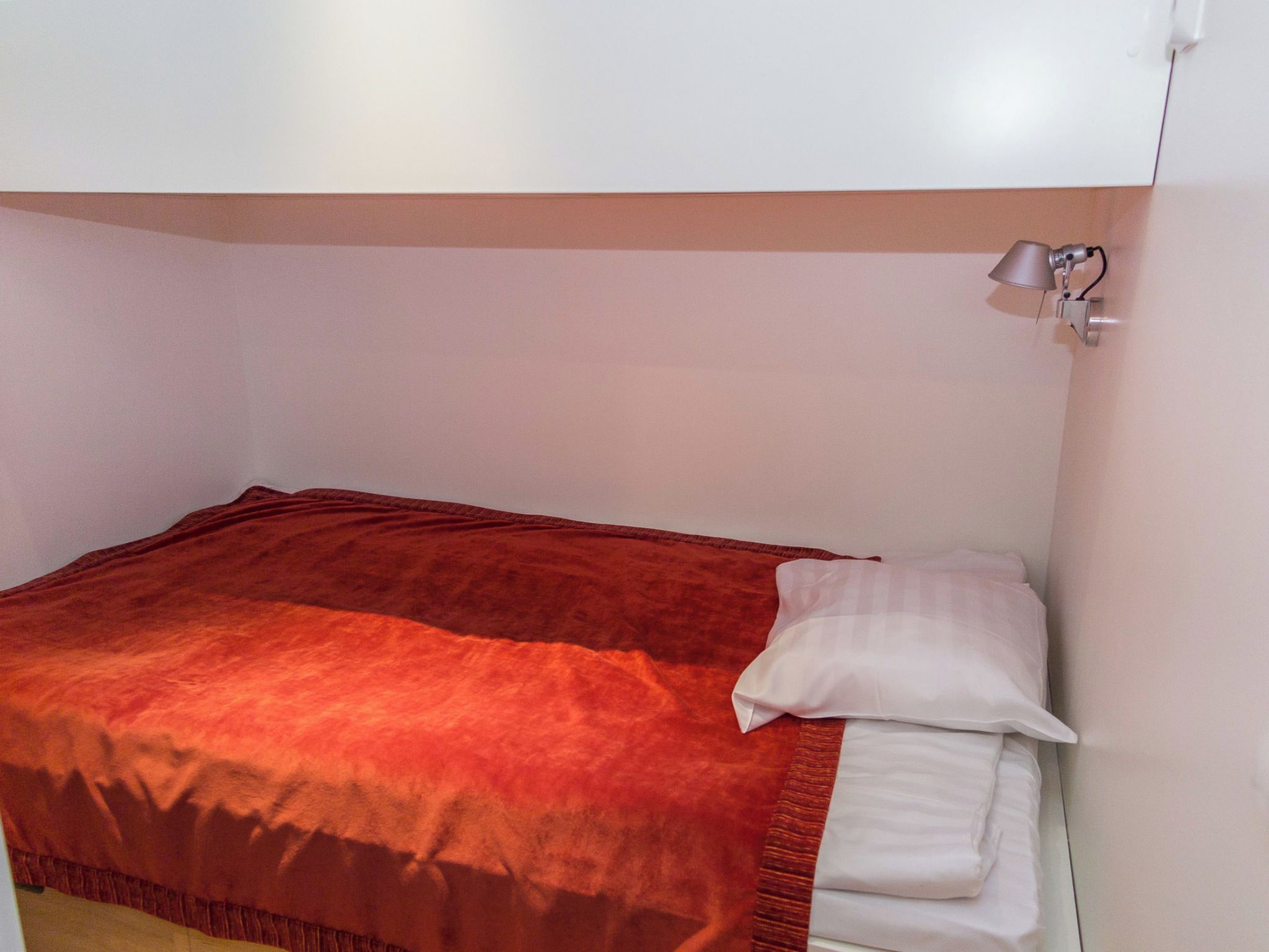 Ein Einzelbett mit roter Decke und weißen Kissen in einem Zimmer mit weißen Wänden und einer an der Decke befestigten Lampe.