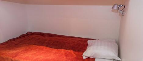 Ein Einzelbett mit roter Decke und weißen Kissen in einem Zimmer mit weißen Wänden und einer an der Decke befestigten Lampe.