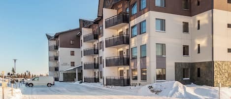Un edificio residencial de varios pisos con balcones, un estacionamiento techado y suelo cubierto de nieve.
