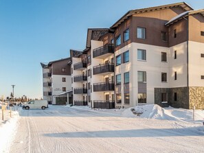 Un immeuble résidentiel à plusieurs étages, doté de balcons, d’un espace de stationnement couvert et d’un sol recouvert de neige.