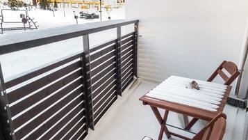 Varanda com mesa e cadeiras de madeira, guarda-corpo de metal e vista para um edifício com varandas.