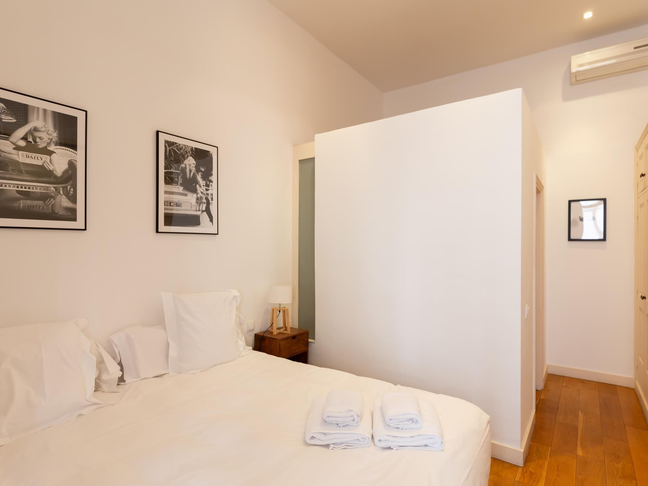 A hotel room with a bed, two framed photographs on the wall, a bedside table with a lamp, and a door leading to another room.