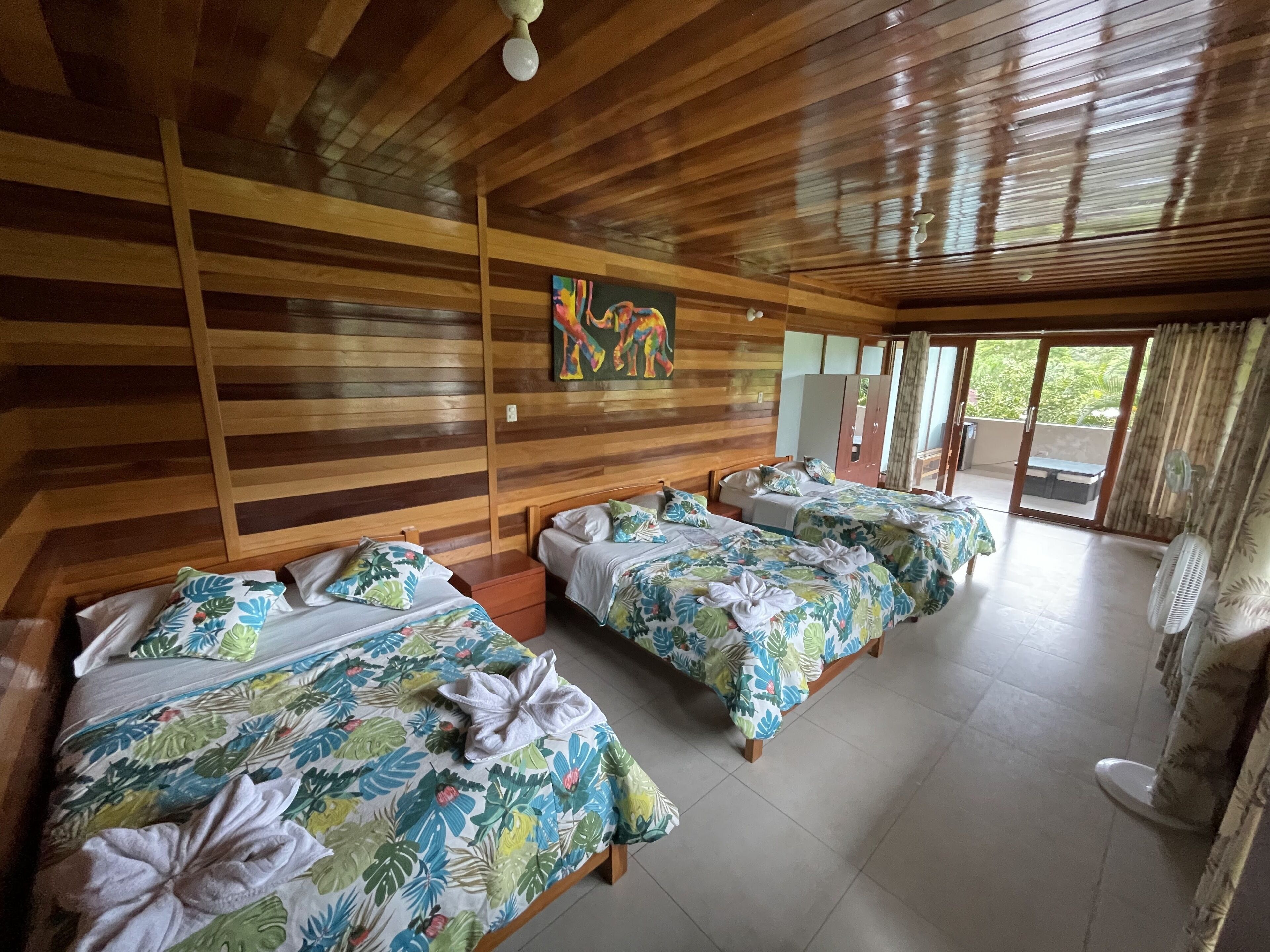 A hotel room with wooden paneling, two beds with floral bedding, a ceiling fan, and a view of greenery through the windows.