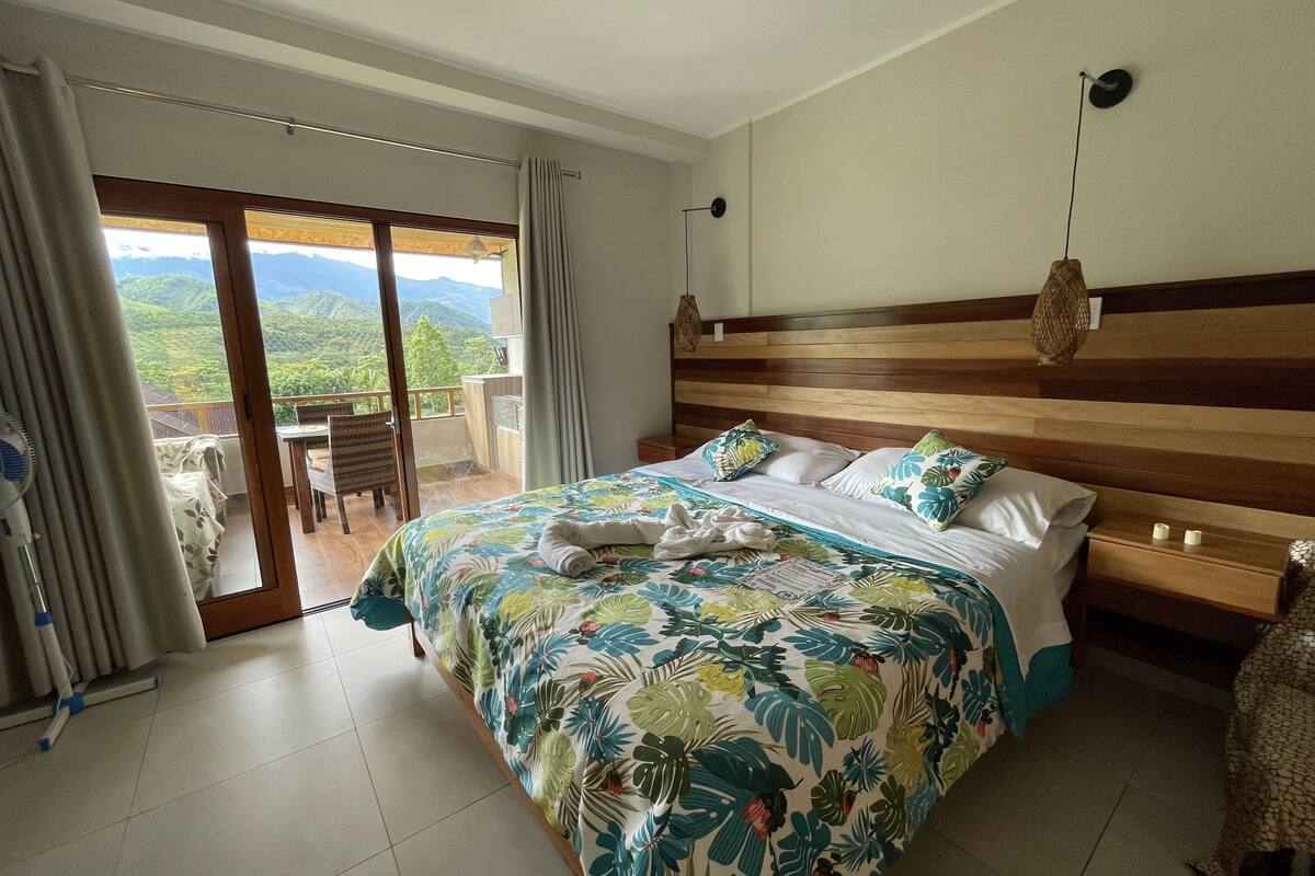 Une chambre d’hôtel dotée d’un grand lit, d’une tête de lit en bois, d’un balcon offrant une vue sur les montagnes et d’un coin repas avec une table et des chaises.