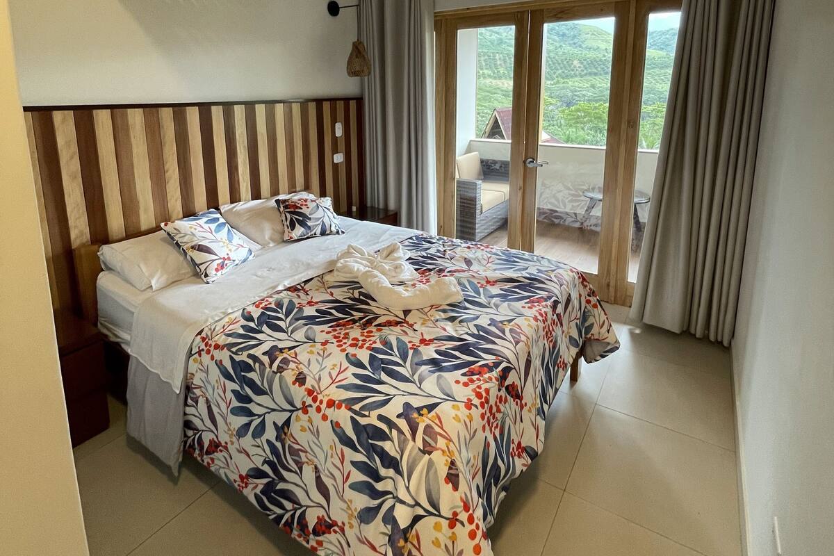 Une chambre à coucher avec un lit, une tête de lit en bois, une fenêtre avec des rideaux et une vue sur la verdure à l’extérieur.