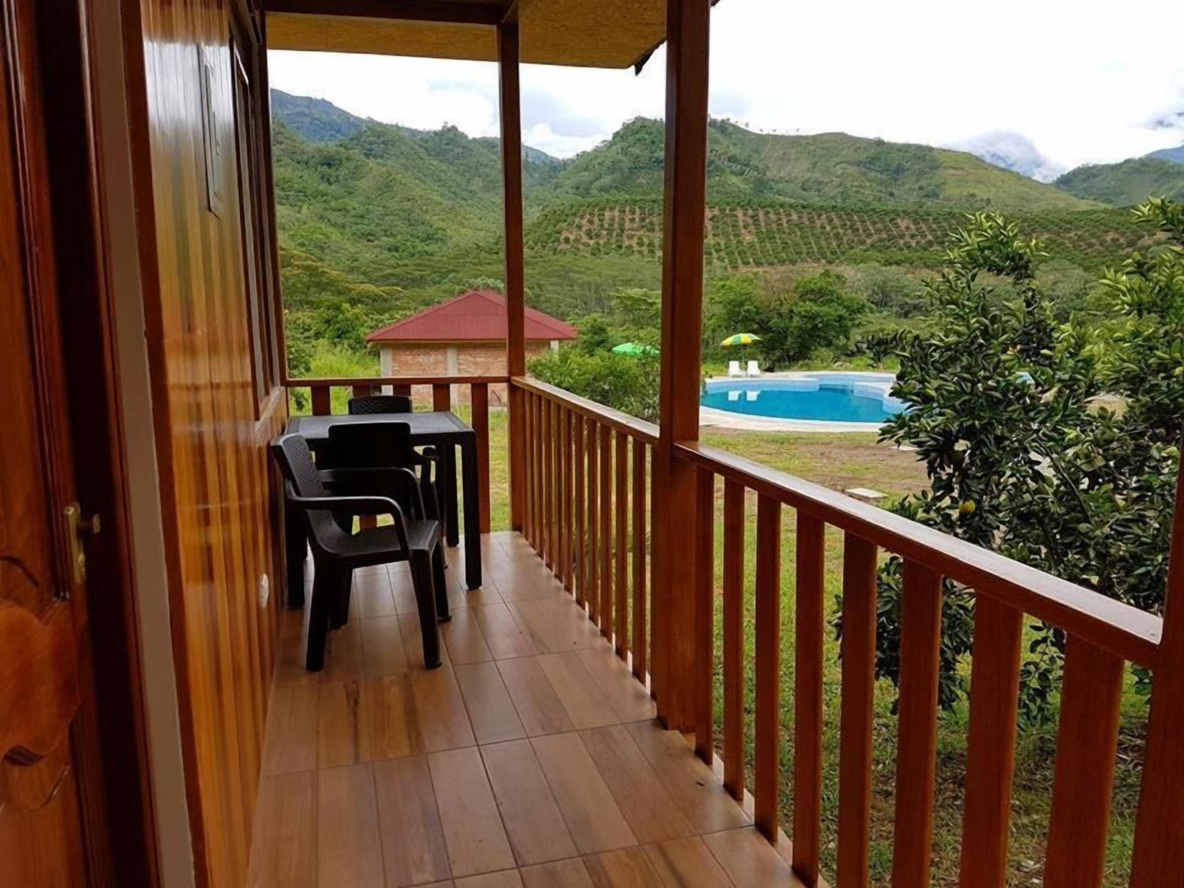A wooden balcony with a table and chairs, overlooking a pool and lush green hills.