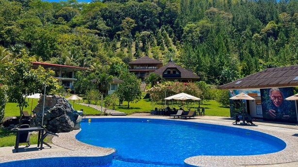 Una piscina con sillas de descanso y sombrillas, rodeada de abundante vegetación y un telón de fondo montañoso.