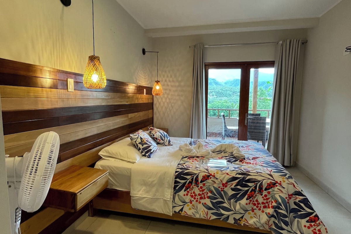 Une chambre avec une tête de lit en bois, un lit recouvert d’une couvre-lit à motifs floraux, un ventilateur sur pied et une vue sur la verdure par la fenêtre.