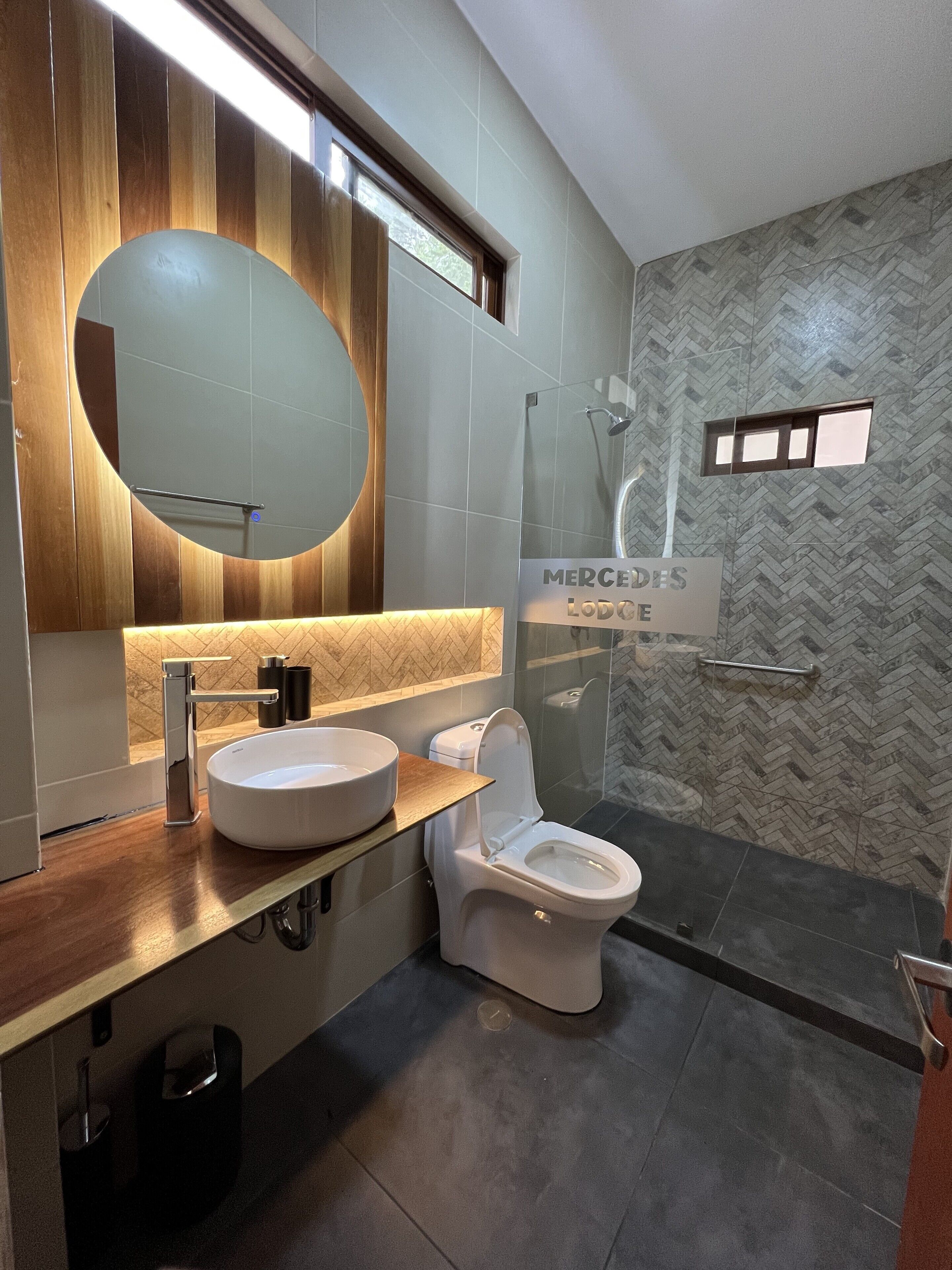 A modern bathroom with a wooden vanity, a round mirror, a toilet, and a shower area.