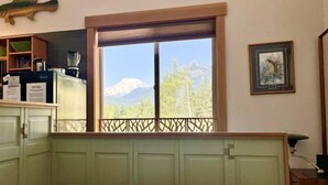 A bedroom with a large window offering a mountain view, a bed, a wooden shelf with kitchen items, and a framed picture on the wall. - Little Big Fish (Packwood)