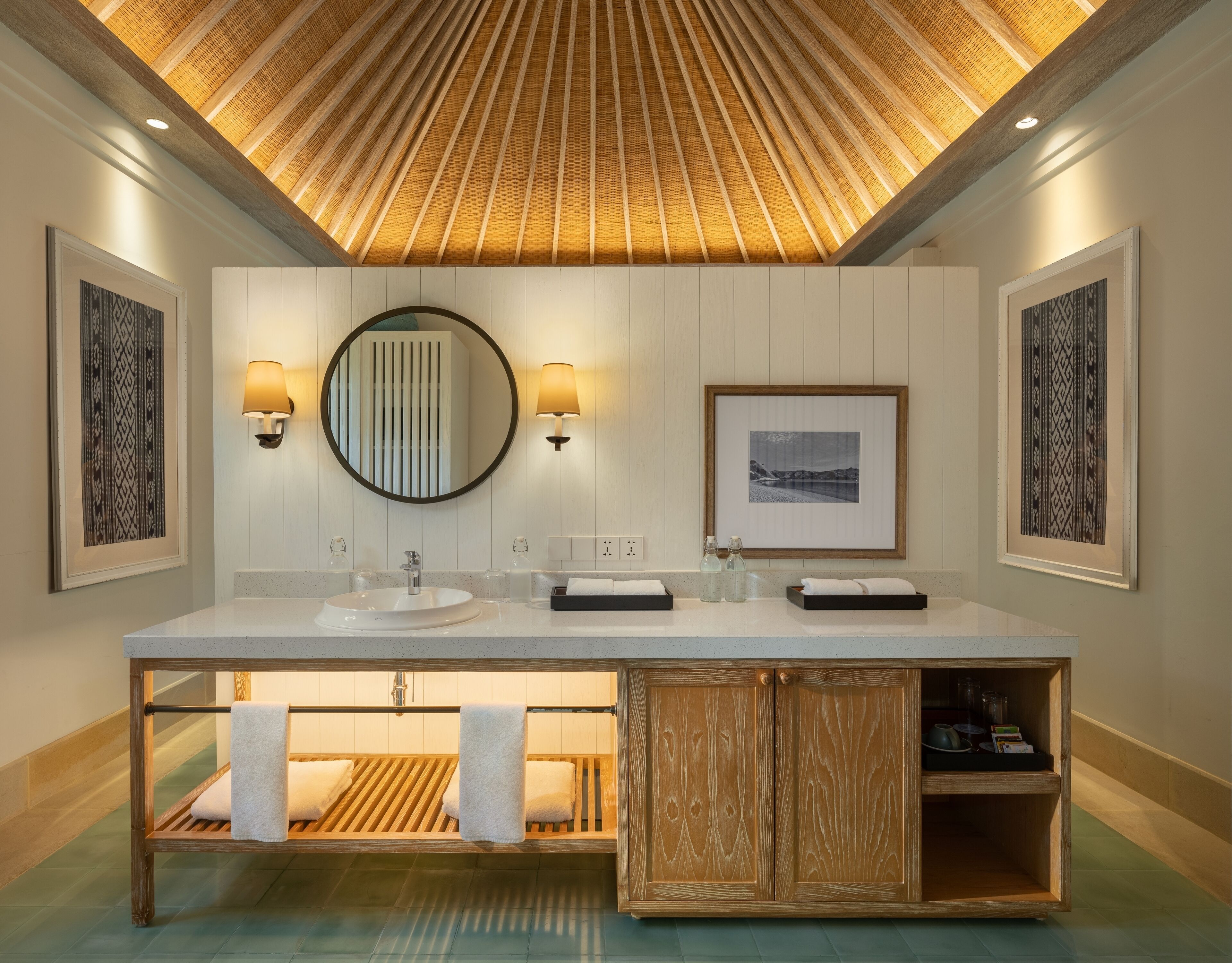 A modern bathroom with a large vanity, a wooden bench with towels, and framed artwork on the walls.