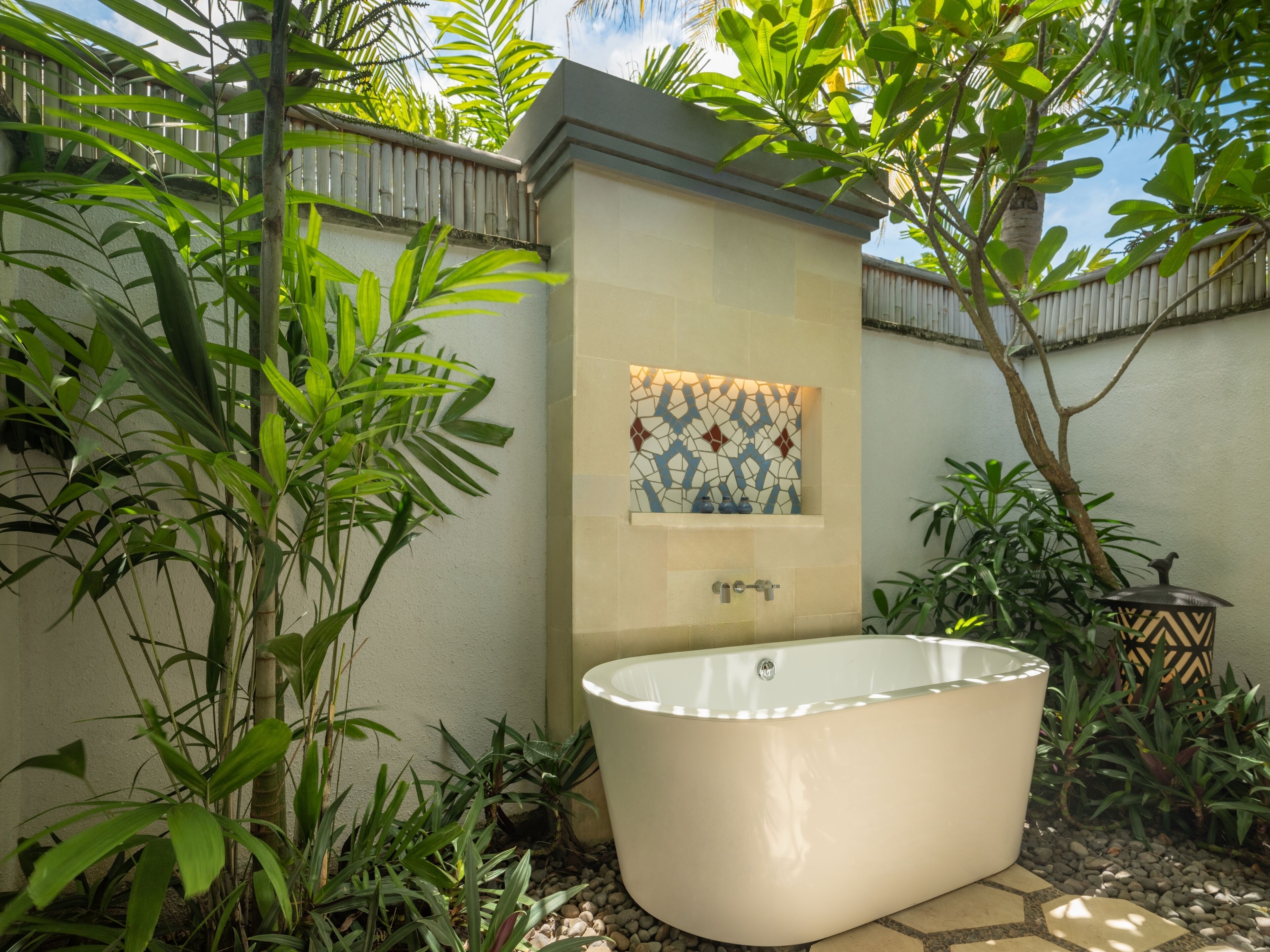 A freestanding bathtub in a garden setting with a tiled wall and a decorative tile above it.
