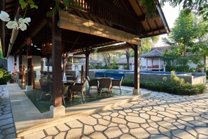 An outdoor patio with a pool, covered seating area, and stone flooring.